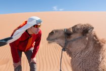 Touriste avec turban et lunettes de soleil embrassant un chameau dans les dunes du désert — Photo de stock