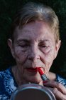 Mulher velha pintando seus lábios em vermelho olhando para si mesma no espelho — Fotografia de Stock