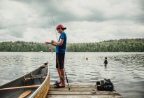 Adolescent garçon pêche à partir d'un quai sur un lac avec des frères nager à proximité. — Photo de stock