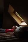 Young boy laying on bed looking up through a sky light in a dark room. — Stock Photo
