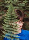 Portrait of young smiling boy covering half his face with fern leaf. — Stock Photo