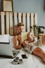 Um homem lê um livro enquanto trabalha com seu celular e laptop em uma cama de hotel. relaxe conceito — Fotografia de Stock