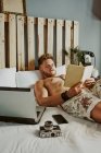 Мужчина читает книгу, когда работает со своим мобильным телефоном и ноутбуком на кровати отеля. Концепция расслабления — стоковое фото