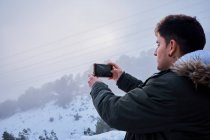 Молодий латинський чоловік фотографує зі своїм телефоном, оточеним снігом — стокове фото