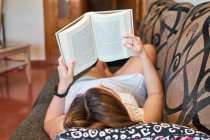 Giovane donna legge un libro sdraiato su un divano — Foto stock
