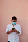 Junger lateinischer Mann mit Maske schaut sein Handy auf rosa Hintergrund — Stockfoto