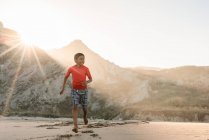 Latino tween chico corriendo en playa con montañas en fondo - foto de stock