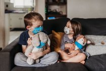 Niña en edad preescolar y niño en edad escolar con máscaras jugando juguetes en el sofá - foto de stock