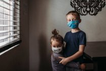 Lifestyle-Porträt junger Geschwister mit Masken beim Umarmen zu Hause — Stockfoto