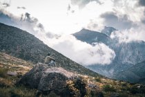 Junger Mann sitzt auf einem Felsen und bewundert Berge mit niedrigen Wolken — Stockfoto