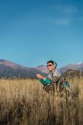 Молодий чоловік з сонцезахисними окулярами сидить у полі, дивлячись на гори дотепно — стокове фото