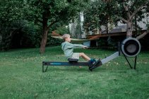 Boy using the rowing machine in his garden at home exercising — Stock Photo