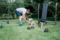 Hombre sosteniendo la pata de sus perros mientras hace ejercicio en casa en cuarentena - foto de stock