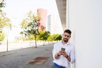 Молодий бородатий чоловік спирається на білу стіну за допомогою мобільного телефону — стокове фото