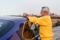 Seitenansicht eines älteren Mannes, der sich auf dem Dach des Fahrzeugs lehnt und bei einer Autofahrt im Grünen nach vorne schaut — Stockfoto