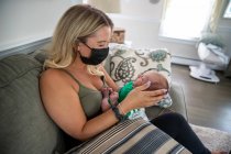 Mulher loira usando máscara facial segurando bebê recém-nascido sonolento. — Fotografia de Stock