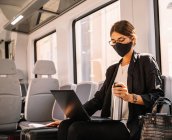 Gerente femenina en mascarilla facial sosteniendo termos y navegando por el portátil mientras viaja al trabajo en tren durante la pandemia - foto de stock