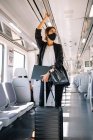 Joven empresaria con mascarilla facial llevando carpeta y bolso y agarrando el manillar mientras viaja al trabajo en tren - foto de stock