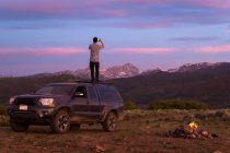 Visão traseira do homem fotografando em pé no carro off-road durante o pôr do sol — Fotografia de Stock