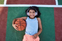 Una niña con el pelo rizado está tirada en una cancha de baloncesto riendo. Concepto de estilo de vida - foto de stock