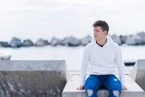 Vista frontal de um adolescente vestindo trajes casuais enquanto sentado em um banco ao ar livre e olhando para longe — Fotografia de Stock