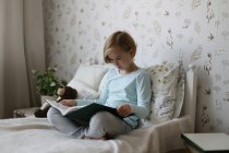 A Russian girl is reading a book on her bed in a bright room. — Stock Photo