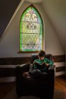 Young boy reading in leather chair in front of ornate window of home. — Stock Photo