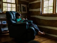 Joven niño leyendo en silla reclinable de cuero en casa cabaña de madera rústica. - foto de stock
