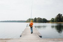Giovane ragazzo che cammina con una canna da pesca su un molo vicino al mare — Foto stock