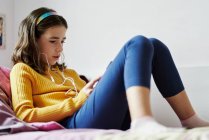 Chica acostada en su cama mirando una tableta y usando auriculares - foto de stock