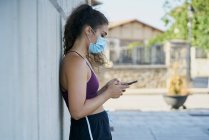 Женщина в спортивной одежде в медицинской маске смотрит на смартфон — стоковое фото