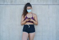 Mujer en ropa deportiva con una máscara médica mirando un teléfono inteligente - foto de stock