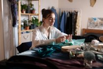 Elegante morena trabajando en casa estudio con pequeñas empresas mientras cose ropa en la mesa - foto de stock