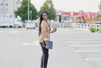 Retrato de mulher negra vestindo um terno marrom segurando um laptop e um recipiente com café andando pela rua — Fotografia de Stock