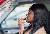 Retrato de uma mulher negra num fato castanho a pintar os lábios dentro do carro. Conceito de negócio e elegância — Fotografia de Stock