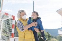Avó usando uma máscara médica com neto em seus braços em um parque no dia ensolarado — Fotografia de Stock