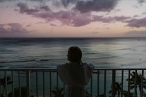Niña en albornoz observa el océano al atardecer desde el balcón en Hawaii - foto de stock