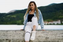 Young woman with blonde braided hair wearing a denim jacket and white jean resting on a bridge on a rainy day — Stock Photo