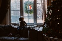 Ein kleines Mädchen sitzt neben einem Weihnachtsbaum und blickt aus dem Fenster. — Stockfoto