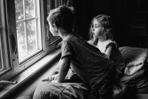Two children perch on a couch and look out a window. — Stock Photo