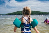 Una niña pequeña con un dispositivo de flotación mira hacia el lago. - foto de stock