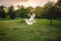 Танцовщица в длинном белом платье кружится в золотом свете в парке на закате — стоковое фото
