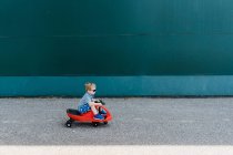 Un ragazzino cavalca una macchina al plasma rossa davanti a un muro verde. — Foto stock