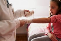Una doctora vendando el brazo de una niña en su habitación. Concepto médico casero - foto de stock