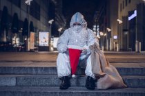 Photo of a beggar, dressed in the coverall used against the coronavirus, the mask, and the beard and hat of Santa Claus, sitting in the stairs in an empty square. Look inside the empty hat. — Stock Photo