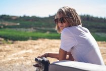Portrait of a girl, with short blonde hair and sunglasses. She looks at the camera, hinting at a smile, while stroking the dog. Sitting on an old sofa, outside, in a rural house. — Stock Photo