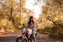 Улыбающаяся молодая женщина, сидящая на мотоцикле на проселочной дороге среди деревьев — стоковое фото