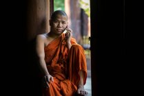 Monje budista vestido con bata naranja llama por un teléfono móvil mientras está sentado en una ventana, Mandalay, Myanmar - foto de stock