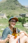 Femme voyageuse trinquant à la bière avec un homme anonyme assis au bord d'un lac dans les montagnes — Photo de stock