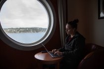 Uma jovem mulher digitando em seu laptop sentado ao lado de uma janela na água — Fotografia de Stock